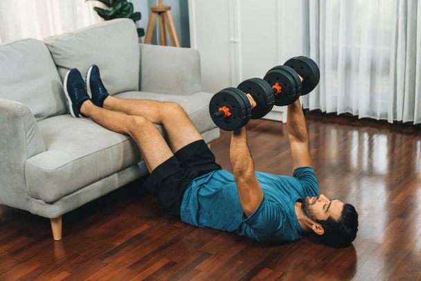 Athletic body and active sporty man using furniture for effective targeting muscle gain with weight lifting dumbbell exercise at gaiety home as concept of healthy fit body home workout lifestyle. - Photo, Image