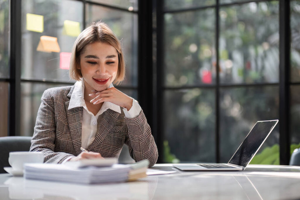 Sorrindo mulher de negócios asiática usa laptop para trabalhar em transações financeiras, cálculos fiscais, planejamento na mesa de trabalho no escritório de vidro.. - Foto, Imagem