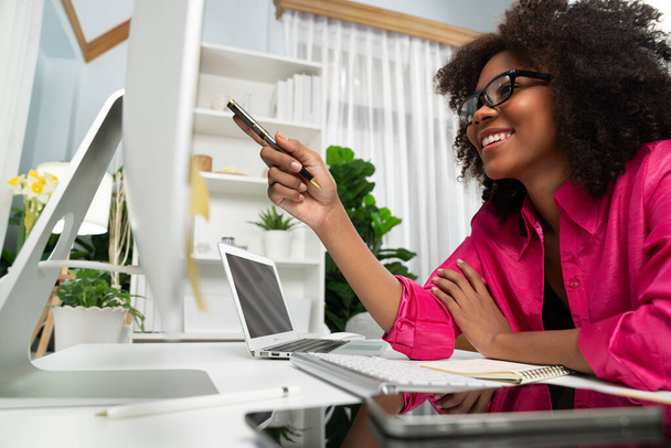 Młoda afrykańska bizneswoman koncentrując projekt na ekranie laptopa monitora, badania centrum danych informacji na stronie komputera i smartfona. Temat miejsca pracy w wygodnym pokoju o czystym kolorze. Tastemaker. - Zdjęcie, obraz