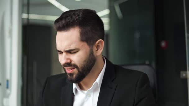 Close up of bearded caucasian man suffering from migraine and touching head while working at personal workplace. Overworked businessman in black suit needing break from work in modern cabinet. - Footage, Video