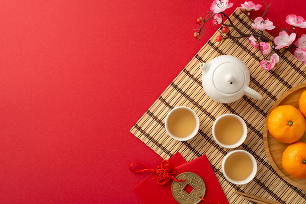 Elegancia tradicional china de año nuevo: imagen de vista superior que muestra lo esencial de la ceremonia del té verde, palillos, mantel de bambú, colgante de moneda simbólica, mandarina, sakura en la superficie roja con espacio para el texto - Foto, Imagen