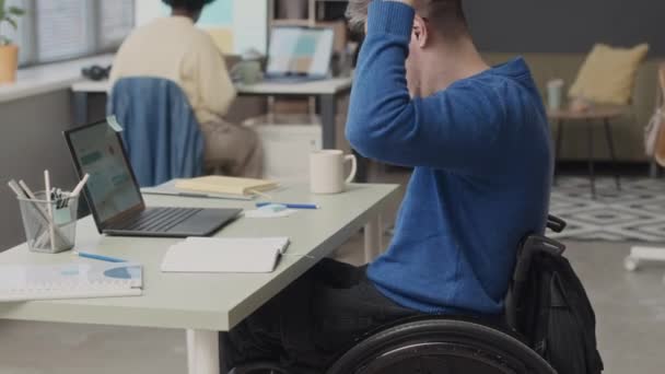 Μέτριο πλάνο του νεαρού Καυκάσου με αναπηρία που κάθεται σε αναπηρικό καροτσάκι στο γραφείο, φορώντας ασύρματα ακουστικά και χρησιμοποιώντας φορητό υπολογιστή, ενώ εργάζεται στο σύγχρονο γραφείο με τη μαύρη γυναίκα συνάδελφο - Πλάνα, βίντεο
