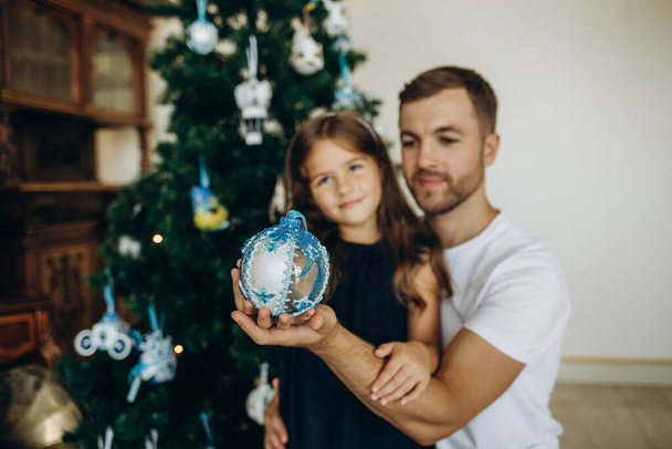 Ο πατέρας δίνει ένα δώρο στην ευτυχισμένη κόρη του κοντά στο χριστουγεννιάτικο δέντρο στο σπίτι. - Φωτογραφία, εικόνα