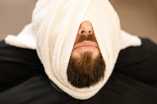A client is lying on a chair in a barbershop with his beard wrapped in a towel. Steaming the beard before the royal shave. - Photo, Image