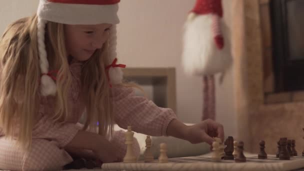 Красивая маленькая девочка в рождественской шляпе и пижаме, играющая в шахматы. - Кадры, видео