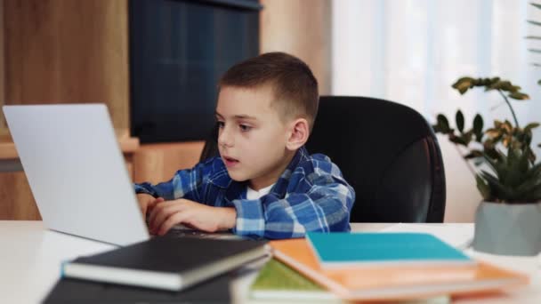 Cute caucasian male child sitting at personal workplace and watching education video on modern laptop. Curious boy looking at camera and using modern gadget to maintaining knowledge outside school. - Footage, Video