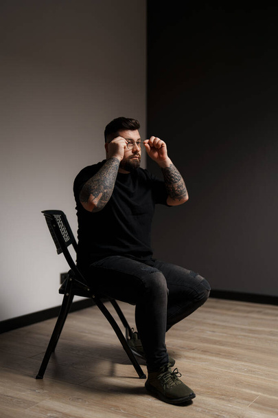 Um homem fashion-forward fixa seus óculos, seus braços tatuados adicionando uma borda à sua pose pensativa - Foto, Imagem