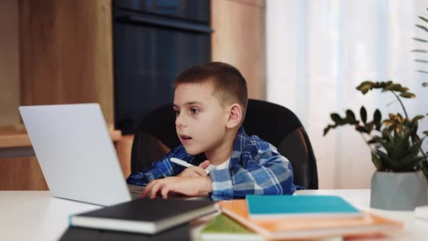 Entzückender kleiner Junge, der Informationen vom drahtlosen Laptop in ein Notizbuch transkribiert, während er drinnen auf einem bequemen schwarzen Stuhl sitzt. Konzentrierter Kaukasier macht Hausaufgaben mit Hilfe moderner Geräte. - Filmmaterial, Video