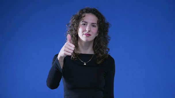 Happy woman showing approval with thumb up, looking at camera while standing on blue background. Approved concept - Photo, Image