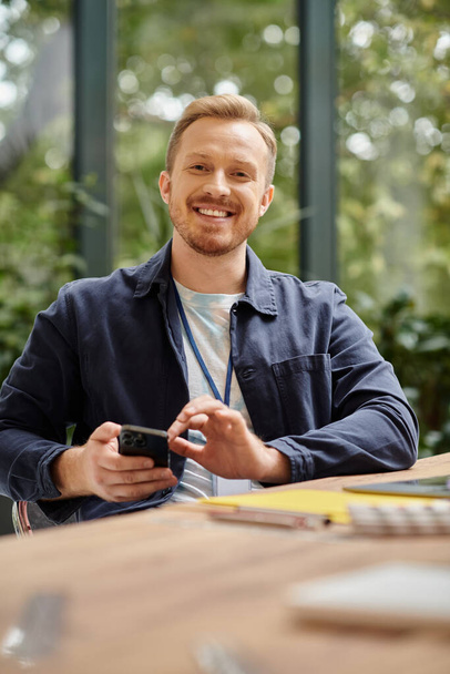 szczęśliwy przystojny mężczyzna w codziennym stroju siedzi przy biurku i pracuje nad jego startupem podczas gdy w biurze - Zdjęcie, obraz