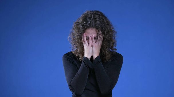 Turhautunut nainen kokee katumusta ja sekaannusta menneistä muistoista, 20-vuotias nainen peittää kasvot häpeässä ja ahdistuksessa seisoen sinisellä taustalla - Valokuva, kuva