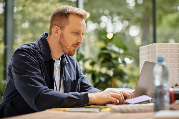 красивый сконцентрированный мужчина в повседневной одежде работает над своим стартапом с ноутбуком в офисе - Фото, изображение