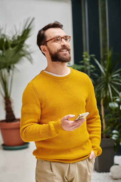 fröhlicher gutaussehender Architekt mit Brille in gelbem Rollkragen posiert mit seinem Handy in seinem Büro - Foto, Bild