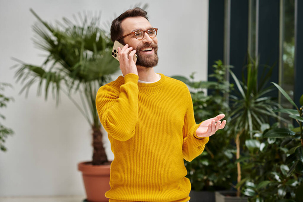 cheerful handsome architect with glasses in yellow turtleneck talking by his phone in his office - Foto, Imagen