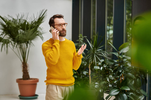 オフィスの携帯電話で話す黄色いタートルネックに集中した魅力的なひげ付き男 - 写真・画像
