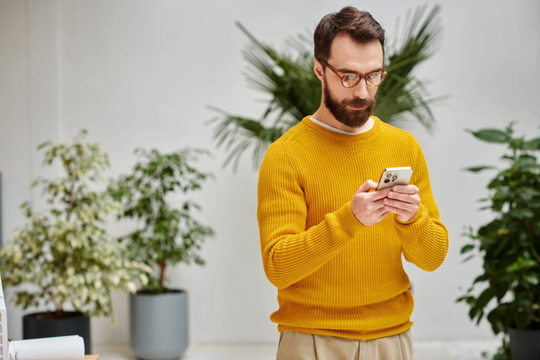 concentrado atraente barbudo homem no amarelo gola alta segurando seu telefone celular no escritório - Foto, Imagem