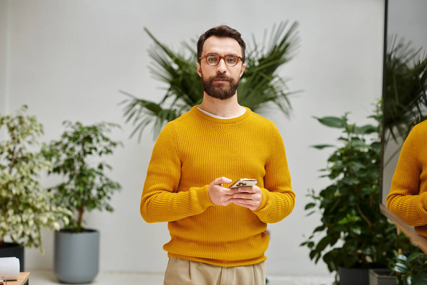Konzentrierter attraktiver bärtiger Mann mit gelbem Rollkragen, Handy in der Hand und Blick in die Kamera - Foto, Bild