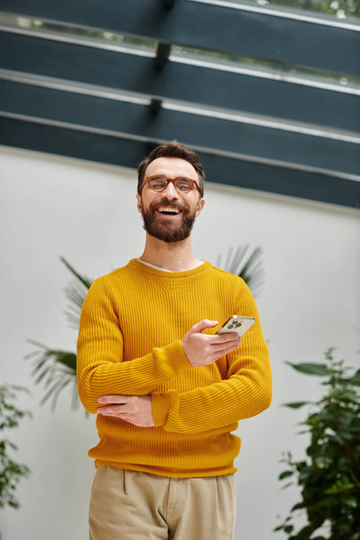 Χαρούμενος όμορφος αρχιτέκτονας με γυαλιά σε κίτρινο ζιβάγκο ποζάροντας με το τηλέφωνό του στο γραφείο του - Φωτογραφία, εικόνα
