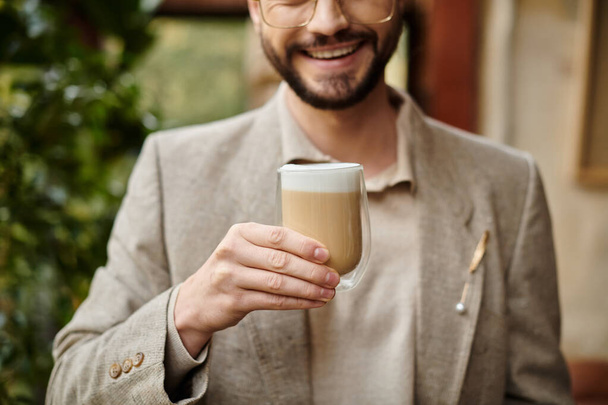 όμορφος ευτυχισμένος άνθρωπος με γενειάδα και συλλέγονται μαλλιά σε κομψό κοστούμι απολαμβάνοντας ζεστό καφέ του - Φωτογραφία, εικόνα