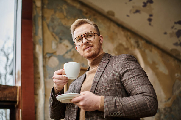 radostný sofistikovaný obchodní vůdce s brýlemi v elegantním elegantním obleku pití jeho horké kávy - Fotografie, Obrázek