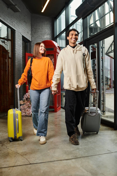 interracial couple heureux marche à travers le hall d'entrée de l'auberge tout en tirant des bagages et en se tenant la main - Photo, image