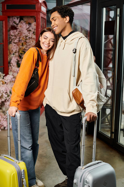 interracial carefree couple standing in hostel entrance hall with colorful suitcases, travel - Photo, Image
