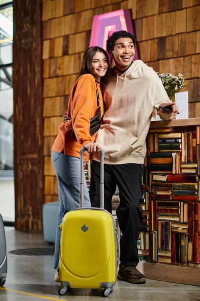 diversa pareja sonriente está junto a su maleta amarilla, vestida con ropa elegante cerca de los libros - Foto, imagen