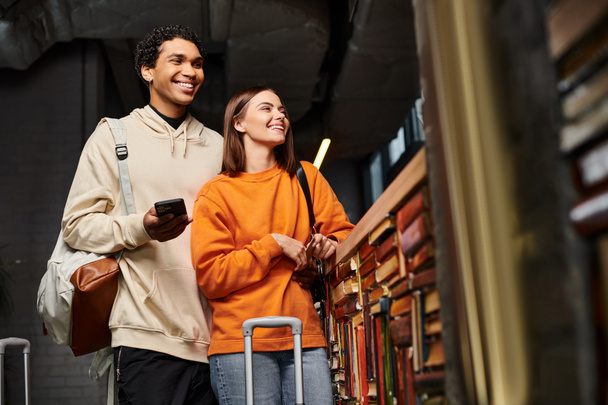 couple heureux et diversifié avec smartphone partageant un moment joyeux dans l'auberge près d'une bibliothèque - Photo, image