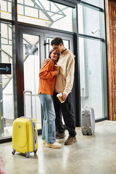 Loving diverse couple with coffee embracing beside travel suitcases in a modern entrance of hostel - Photo, Image