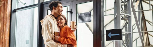 feliz pareja diversa viaje con café abrazando al lado de la entrada de cristal moderno del albergue, pancarta - Foto, Imagen