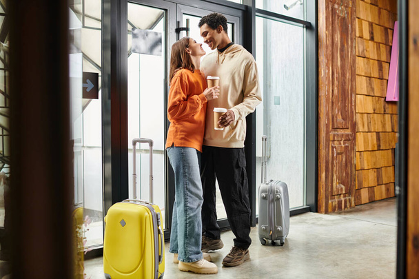 happy interracial couple with coffee embracing beside travel luggage in a modern entrance of hostel - Foto, Imagem