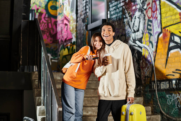diverse couple smiling and standing next to each other on stairs with graffiti, man holding luggage - Photo, Image