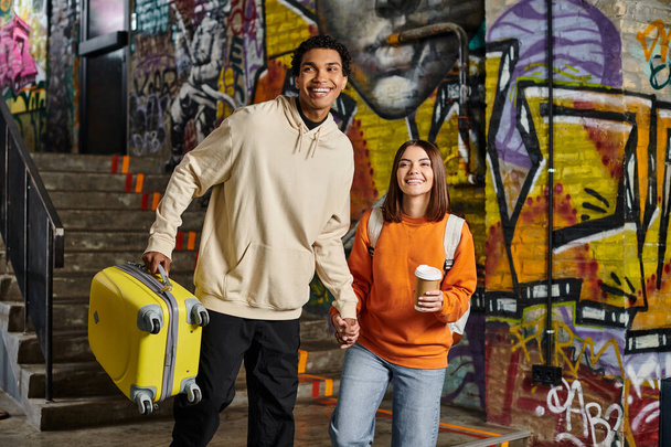 pareja diversa tomados de la mano y sonriendo con un equipaje amarillo en una pared pintada de graffiti, albergue - Foto, Imagen