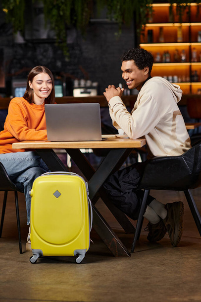 feliz pareja diversa mirando a la computadora portátil, hombre negro y mujer sentado en la mesa cerca de equipaje de viaje - Foto, imagen