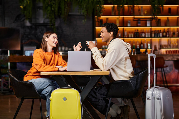 черный мужчина и женщина смеются и разговаривают за столиком кафе, с чемоданами рядом с ними, ноутбук - Фото, изображение