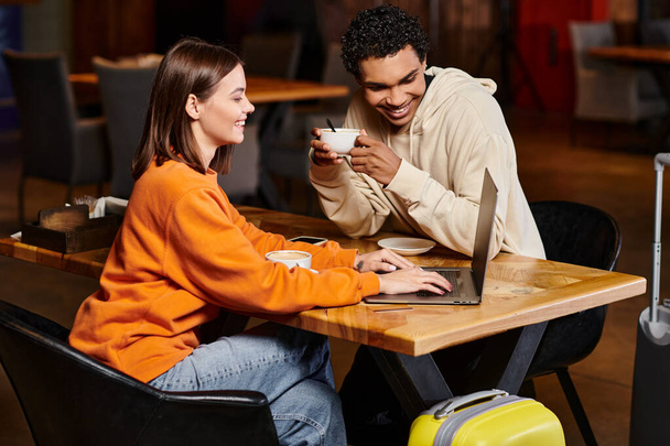 Mujer feliz mostrando algo en el ordenador portátil al hombre negro mientras sostiene la taza de café en la cafetería, diversidad - Foto, imagen