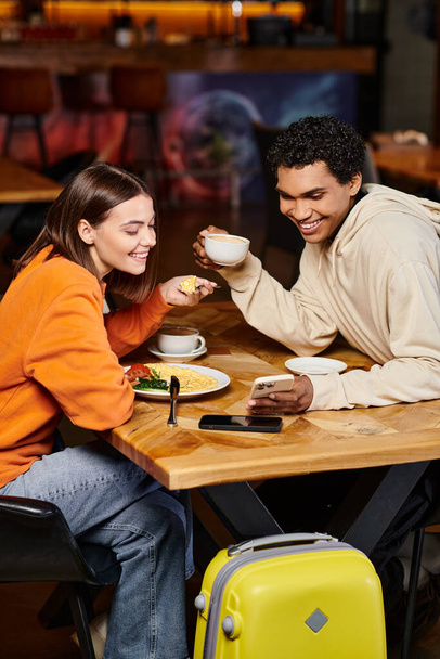 diverse couple looking at smartphone over coffee, black man and pretty woman in hostel cafe - Fotoğraf, Görsel