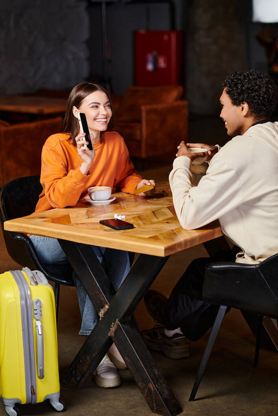 black man and woman using smartphone while sitting at table with coffee in cafe, luggage and travel - Фото, изображение