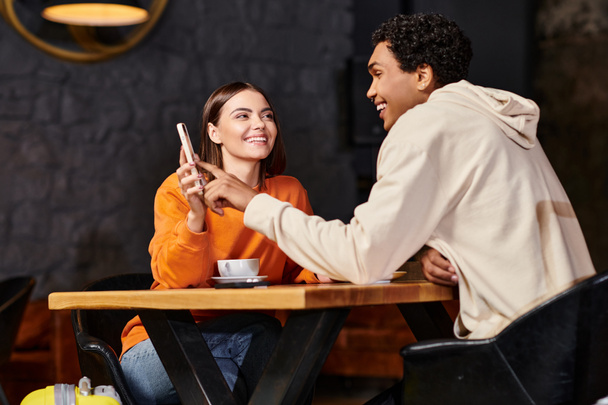 オレンジのセーターの幸せな女性はカフェに座っている間黒いボーイフレンドを笑顔にするために電話を示します - 写真・画像