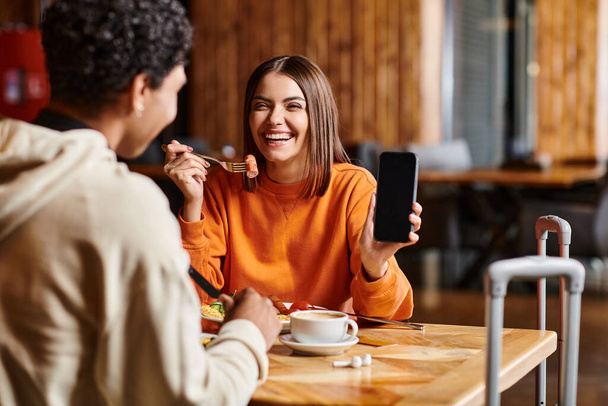 feliz jovem mulher em camisola laranja feliz mostrando seu telefone para o namorado preto durante a refeição - Foto, Imagem