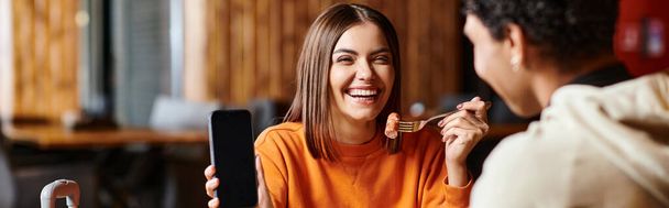mujer feliz en suéter naranja felizmente mostrando su teléfono al novio negro durante la comida, pancarta - Foto, Imagen