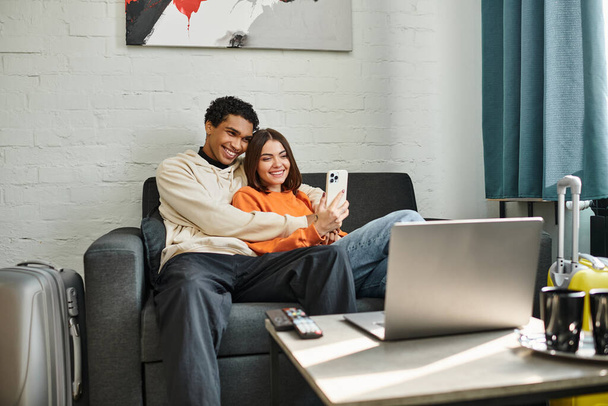 Een paar vangt een moment van liefde en technologie als ze glimlachen voor een selfie in hostel woonkamer - Foto, afbeelding