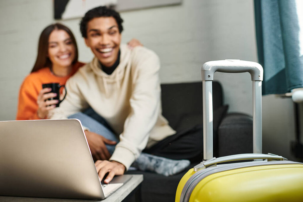 focus on yellow luggage, happy diverse couple traveling together hostel living room and laptop - Valokuva, kuva