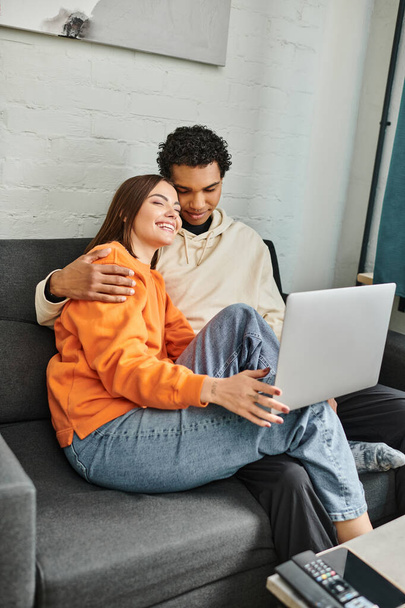 happy diverse couple snuggle on couch while watching movie on laptop, hostel room - Photo, Image