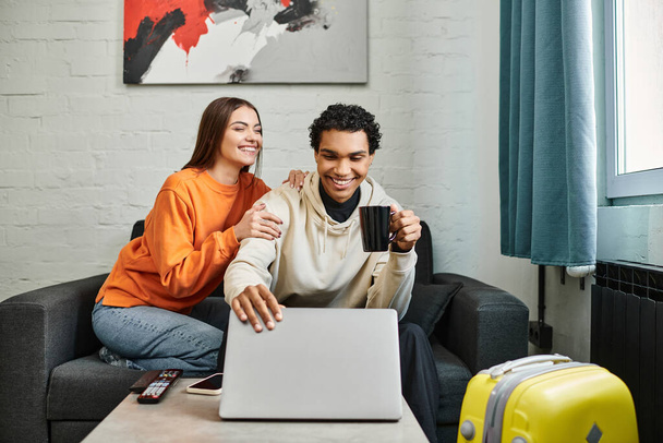 Excited diverse couple seated on couch with a laptop, possibly booking a trip in hostel room - Photo, Image