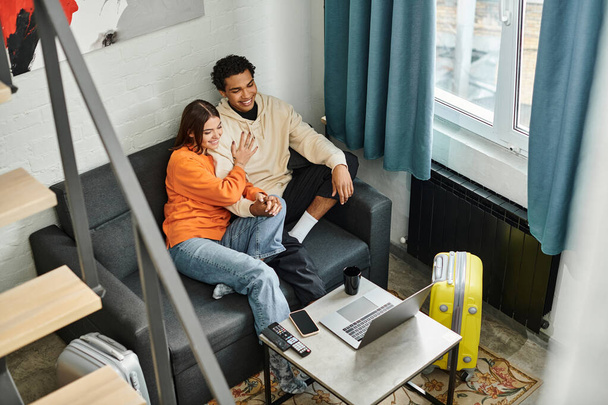 Молодая многонациональная пара наслаждается тихим моментом на уютном диване с ноутбуком в современном хостеле - Фото, изображение