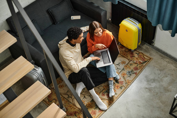 diverse couple looking at a laptop while planning their next trip, sitting on carpet near stairs - Photo, Image