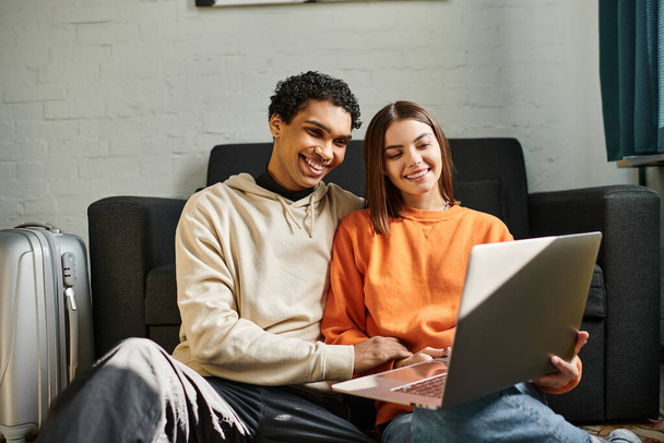 Χαμογελώντας πολυεθνικό ζευγάρι άνετα χρησιμοποιώντας ένα φορητό υπολογιστή μαζί σε ένα σκοτεινό καναπέ, ζευγάρι στόχους - Φωτογραφία, εικόνα