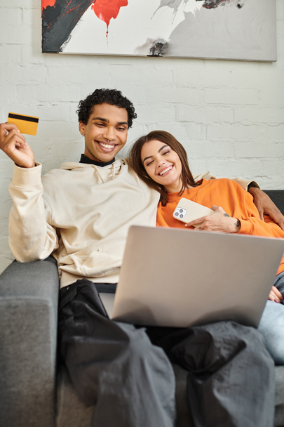 ευτυχισμένο πολυεθνικό ζευγάρι ψώνια σε απευθείας σύνδεση, κρατώντας μια πιστωτική κάρτα, με ένα φορητό υπολογιστή στον καναπέ σε ξενώνα - Φωτογραφία, εικόνα