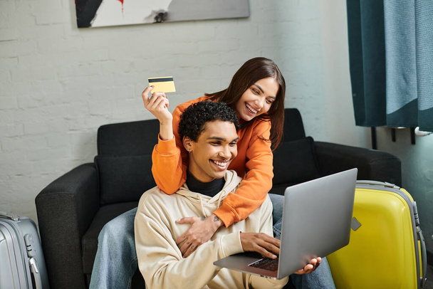 felice coppia multietnica shopping online, in possesso di una carta di credito, con un computer portatile vicino ai bagagli - Foto, immagini